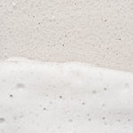 A close-up texture shot of a white and beige speckled surface, showing a horizontal line of thick white foam or soap bubbles across the lower half.