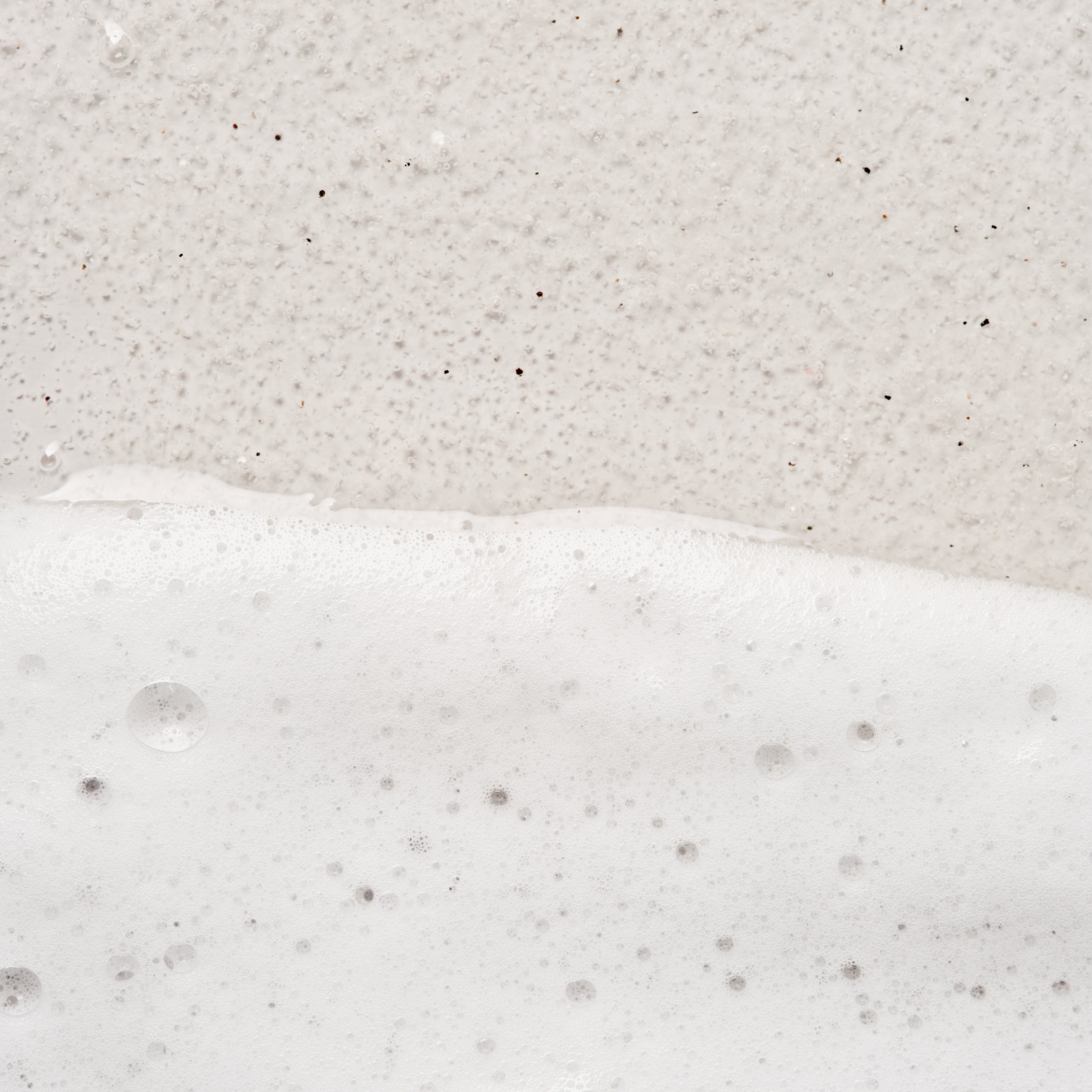 A close-up texture shot of a white and beige speckled surface, showing a horizontal line of thick white foam or soap bubbles across the lower half.